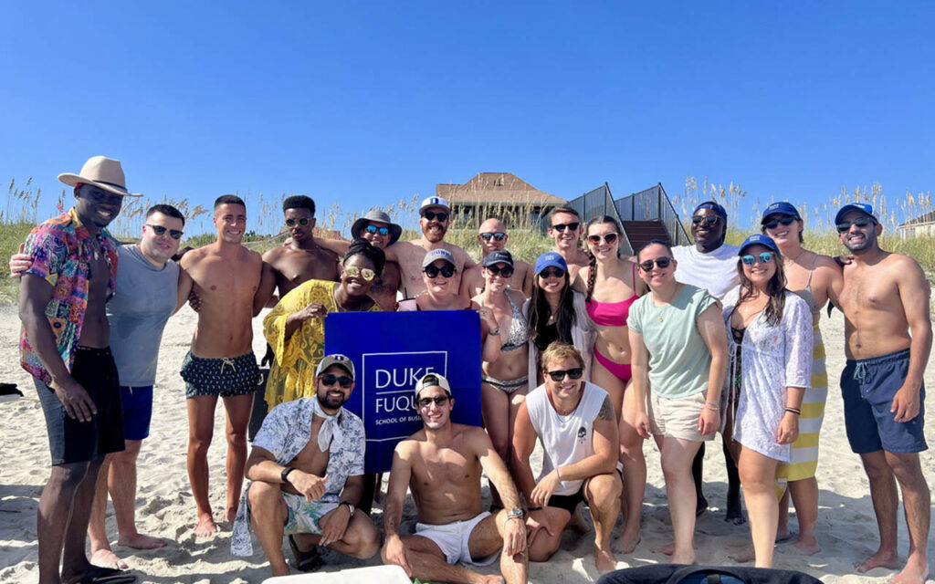 Mayank Tulsiana, Accelerated MBA student and MQM alumnus at Duke University's Fuqua school of Business with a group of Fuqua friends at the beach. They are kneeling and standing around a sign reading "Fuqua School of Business"