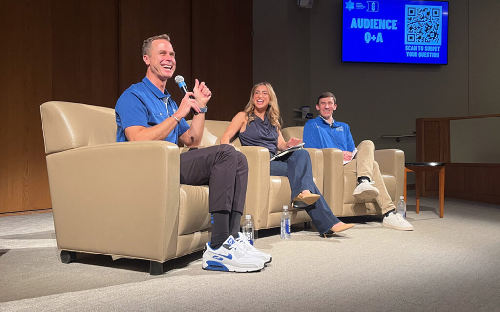 Duke men's basketball coach Jon Scheyer sitting on stage in a tan arm chair, students Sofie Jacobs and Ben Schick sit in identical chairs to the right