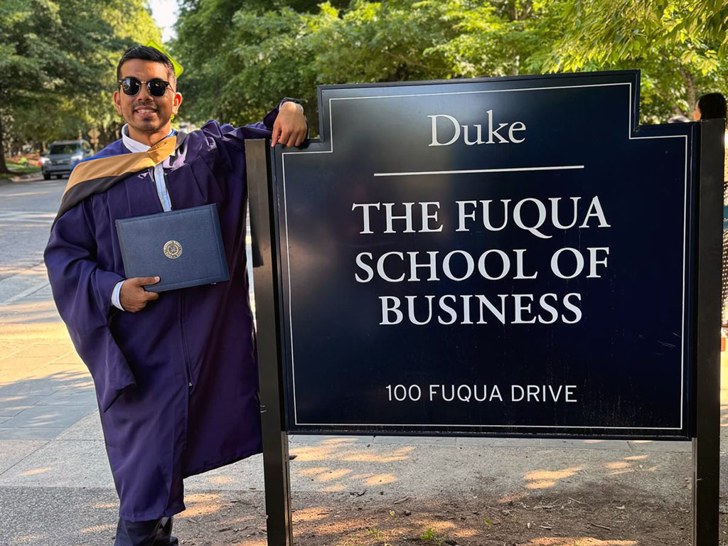 Sai Kuncham Venkat wearing his graduation gown, leans on a sign reading "The Fuqua School of Business"