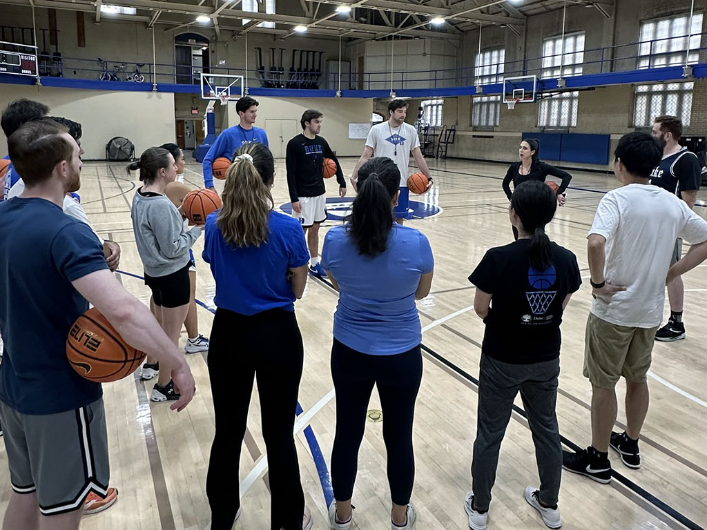 Daytime MBA students standing in a circle, receiving direction from second-year MBA student Stephanie Blair at a Duke gym