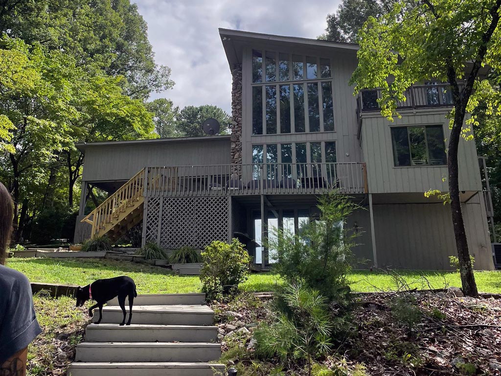 A gray lake house with a wall of windows at the top of a slight hill with about 6 stairs leading up, a black dog is walking about halfway up the stairs