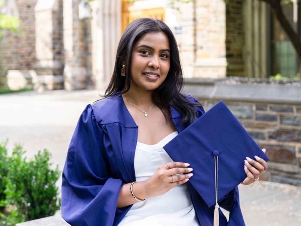 Monika Palabindela wearing a blue gown draped over a white dress. She is holding her graduation cap.