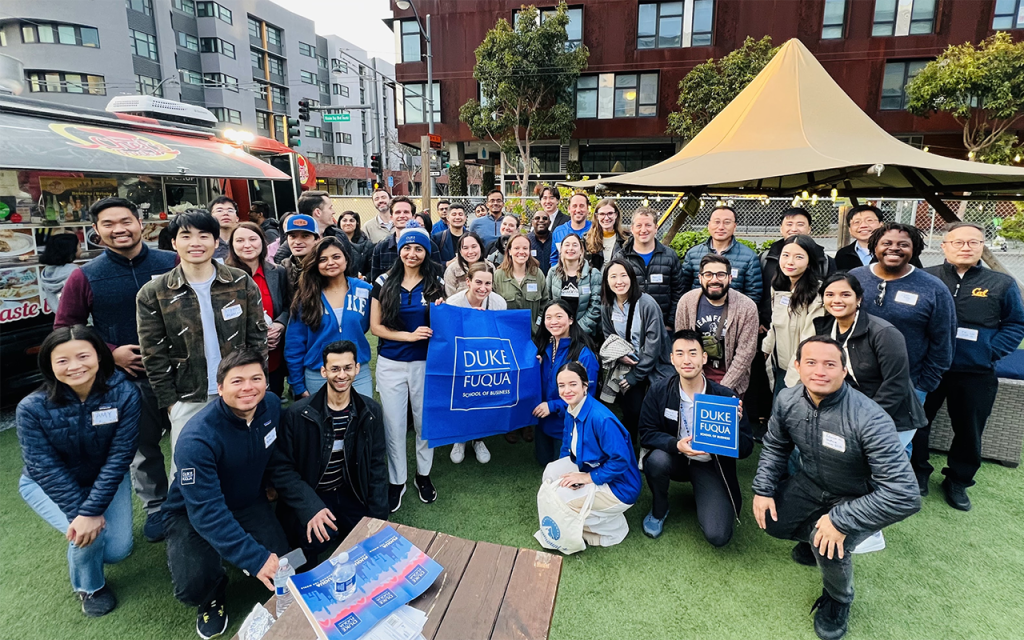 A group of at least two dozen Fuqua alumni post for a photo on a patio in San Francisco. Someone is holding a blue Duke Fuqua flag front and center.