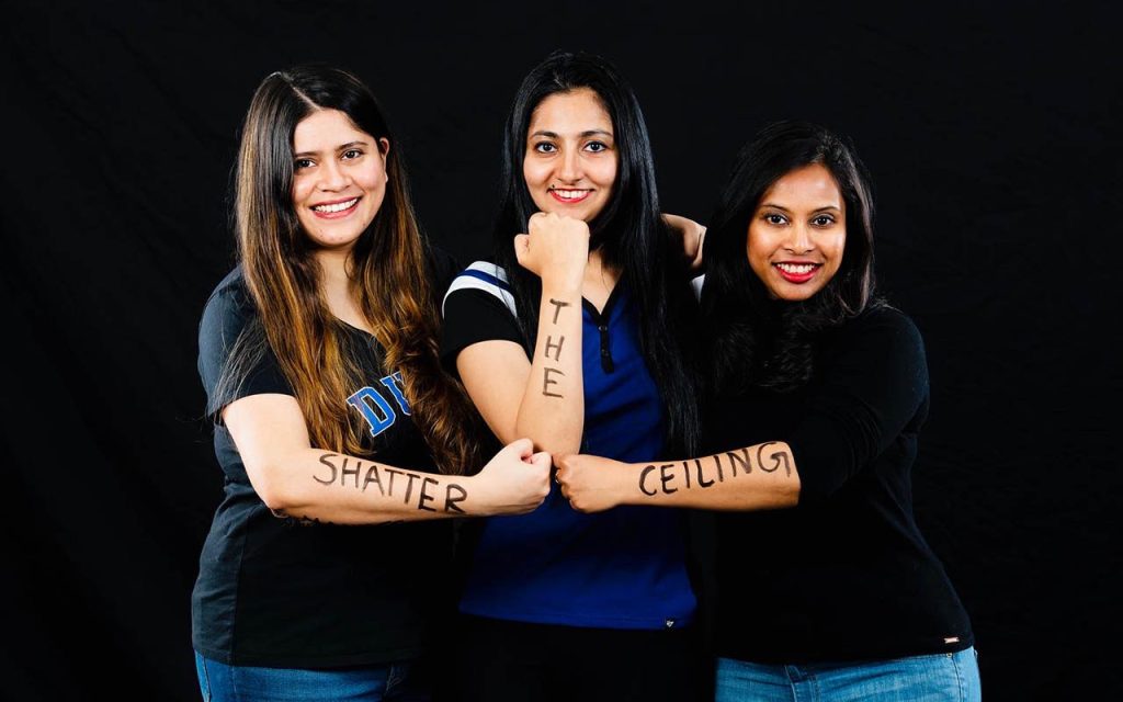 Ayushi Saxena with two friends standing in front of a dark backdrop. They each have words written on their fore arms, reading (from left to right) Shatter the Ceiling