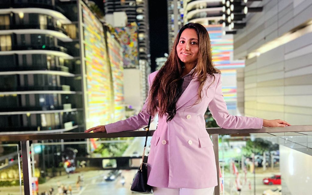 Ayushi Saxena stands on a bridge over a busy street. She is wearing a light-colored pantsuit.