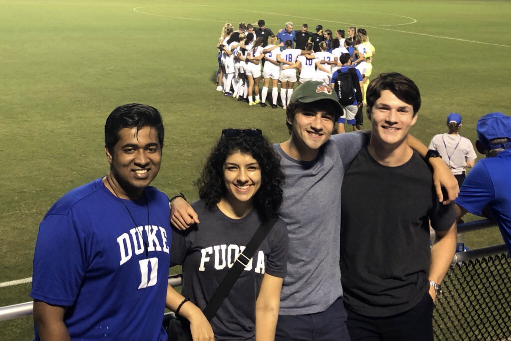 four students posing for a photo with the Duke Women's Soccer student-athletes huddling behind them on the field