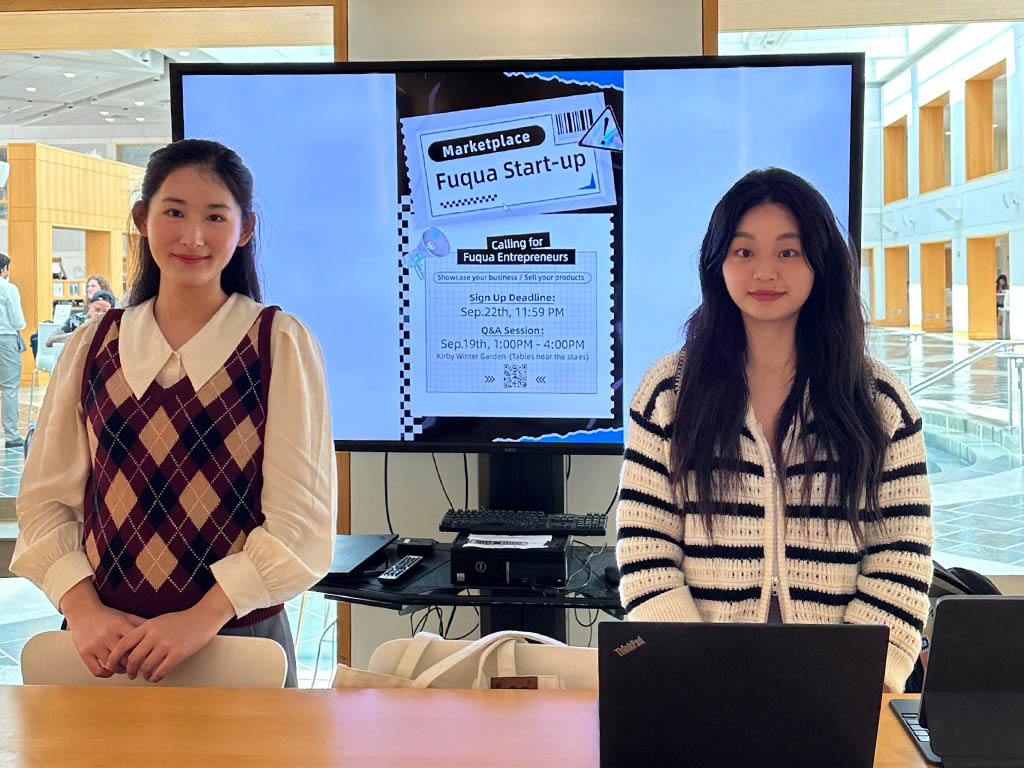 Nolan Gao stands to the right of Jiayi Kong in front of a monitor citing various information of the Fuqua Startup Marketplace