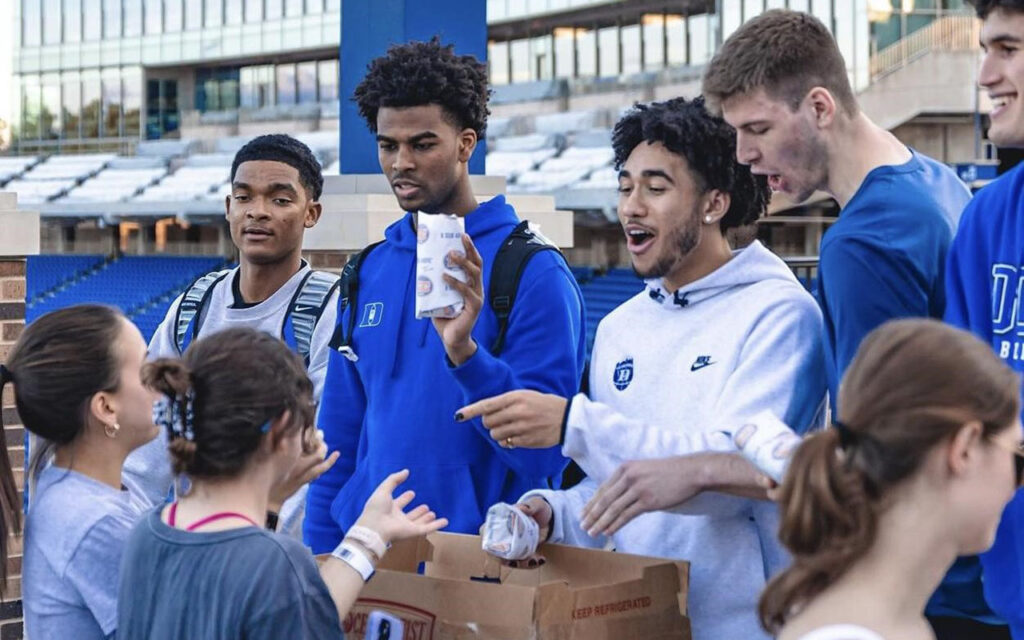 Four men's Duke basketball players handing out packed meals to students camping out ahead of the game