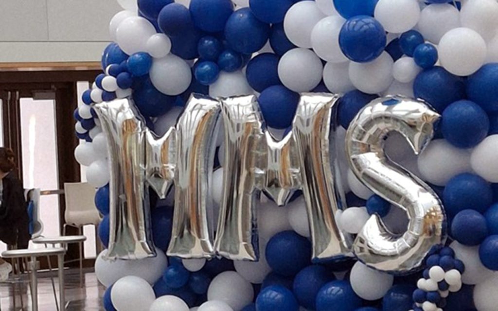 Silver balloons spelling "MMS" in front of a backdrop of blue and white balloons