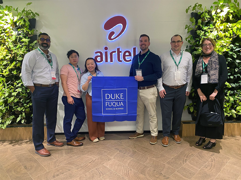 Matt Engle and 5 of his Global Executive MBA classmates standing in front of an Airtel India sign. Matt and a woman are holding a blue flag with the Duke Fuqua logo.