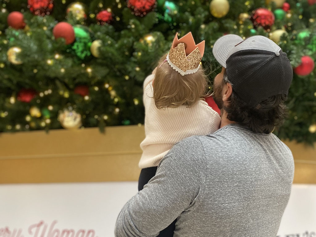 Kyle Goodman, a student in the Weekend Executive MBA program at Duke University's Fuqua School of Business, holding his young daughter, facing away from the camera toward a decorated christmas tree