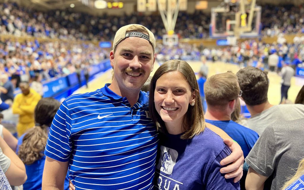 Mary Dohrmann stands to the right of her husband and classmate. They are both wearing Duke apparel in the stands of a Duke basketball game at Cameron Indoor Stadium.