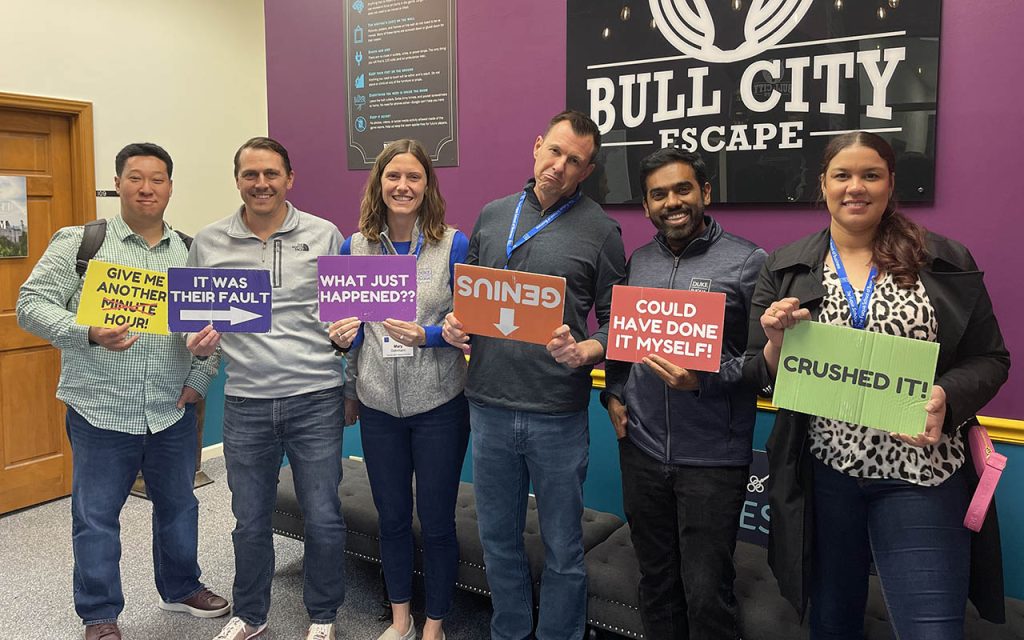 Mary Dohrmann and a few of her Weekend Executive MBA classmates holding signs after completing an escape room, a sign reading "Bull City Escape" is behind them