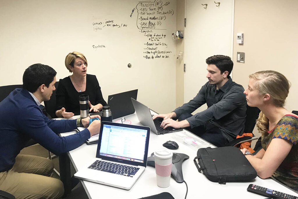 Erin and students in a discussion, seated at a conference table with laptops out and notes about the social impact conference on the white board