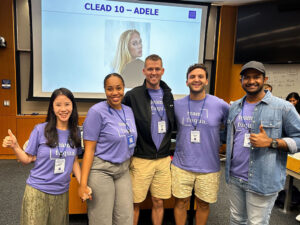 Gerard Pozzi with his teammates wearing the same purple shirt, standing in front of a screen projecting an image of Adele at Duke University's Fuqua School of Business