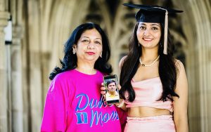 Ayushi Saxena with her mother, Ayushi is wearing a graduation cap and her mother is wearing a pink shirt that reads "Duke Mom." Ayushi is holding a picture of her late father on her phone.