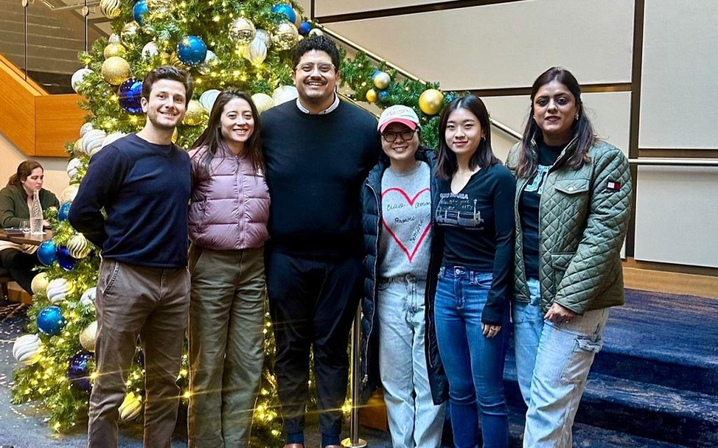 Vertika Srivastava stands to the right of five first-year MBA students she is mentoring as an FCCP Fellow. They are standing at the bottom of the staircase at the J.B. Duke Hotel dining room, which is decorated for Christmas.