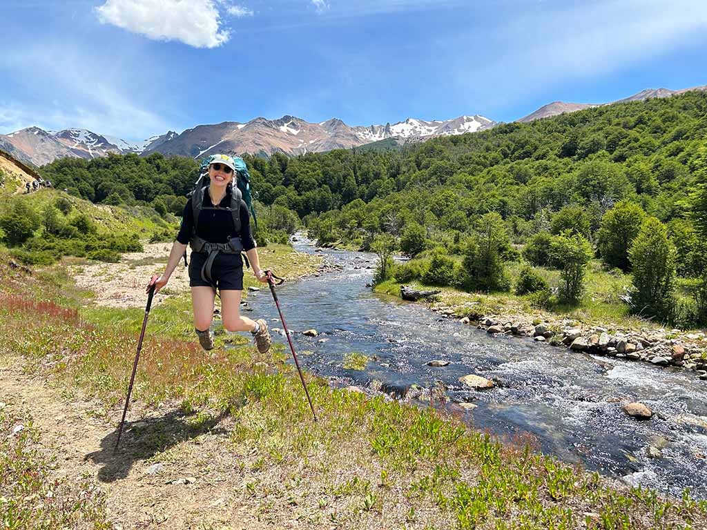 Frannie Gurzenda, a Daytime MBA student at Duke University's Fuqua School of Business, jumps in the air with trekking poles firmly planted on grass near a trail. A flowing river and trees are in the background.