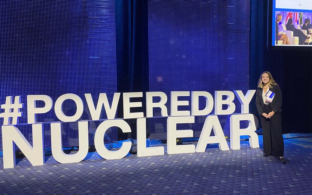 Emma Derr stands beside large “#POWEREDBYNUCLEAR” sign lit up in an indoor event hall