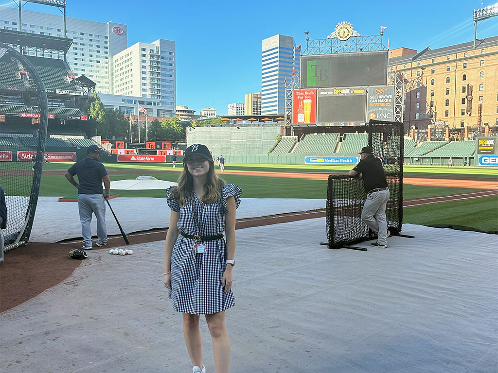 Emma Derr sands on a baseball field near batting practice nets with stadium seating and a scoreboard in the background.