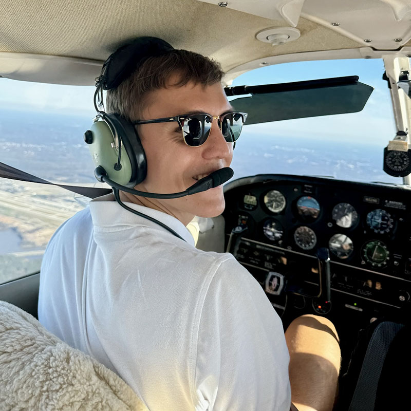 Jan Delkeskamp, a student in the Master of Management Studies program at Duke University's Fuqua School of Business, sits in the pilot seat of a small aircraft in flight. He is wearing a headset and looking back to the second row of the plane.