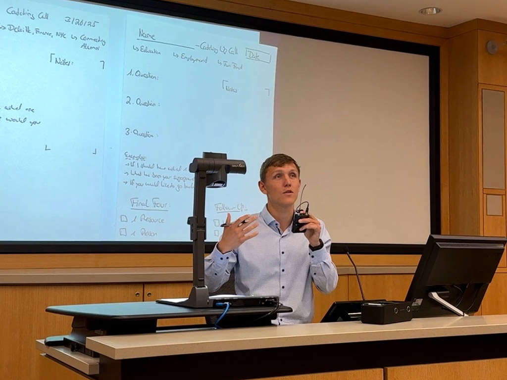 Jan Delkeskamp speaking into a mic and gesturing in front of a projector screen in a classroom at Duke University's Fuqua School of Business
