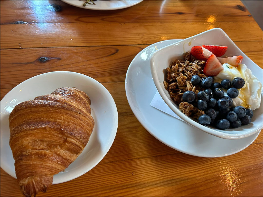 A rustic café table at Guglhupf holds a croissant on one plate and a bowl with granola, yogurt and fruit in another