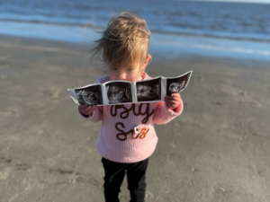 The daughter of Iain Turnbull, a student in the Global Executive MBA Class of 2024, holding ultrasound photos wearing a t-shirt reading "big sis"