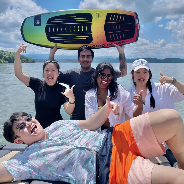 Betty Sun with her MQM capstone classmates on a boat on a river, one is holding a wakeboard over his head, another is laying horizontally in front of the group