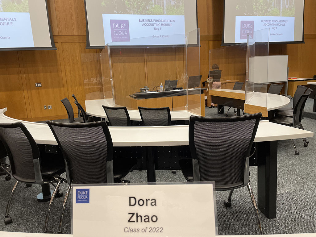 A photo taken from a desk in a classroom at Duke University's Fuqua School of Business. In the foreground, there is a nametag that reads "Dora Zhao, Class of 2022"