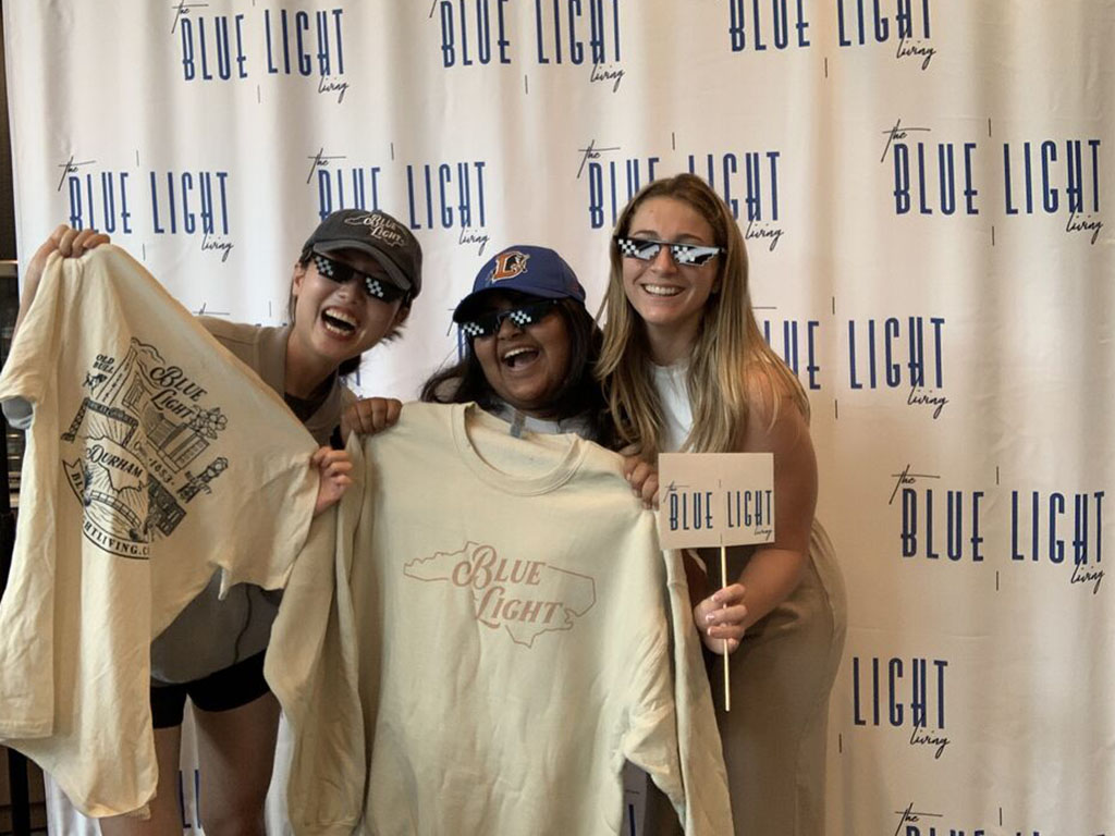 Caterina Mora and her two roomates standing in front of a "Blue Light" backdrop, holding branded flags and t-shirts from the Durham apartment complex near Duke University's Fuqua School of Business.