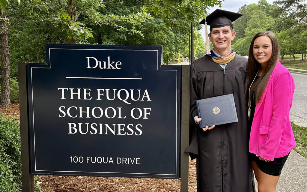 Joshua Duncan wearing a graduation gown and holding a diploma, stands with his wife next to a sign reading, "Duke - The Fuqua School of Business 100 Fuqua Drive"