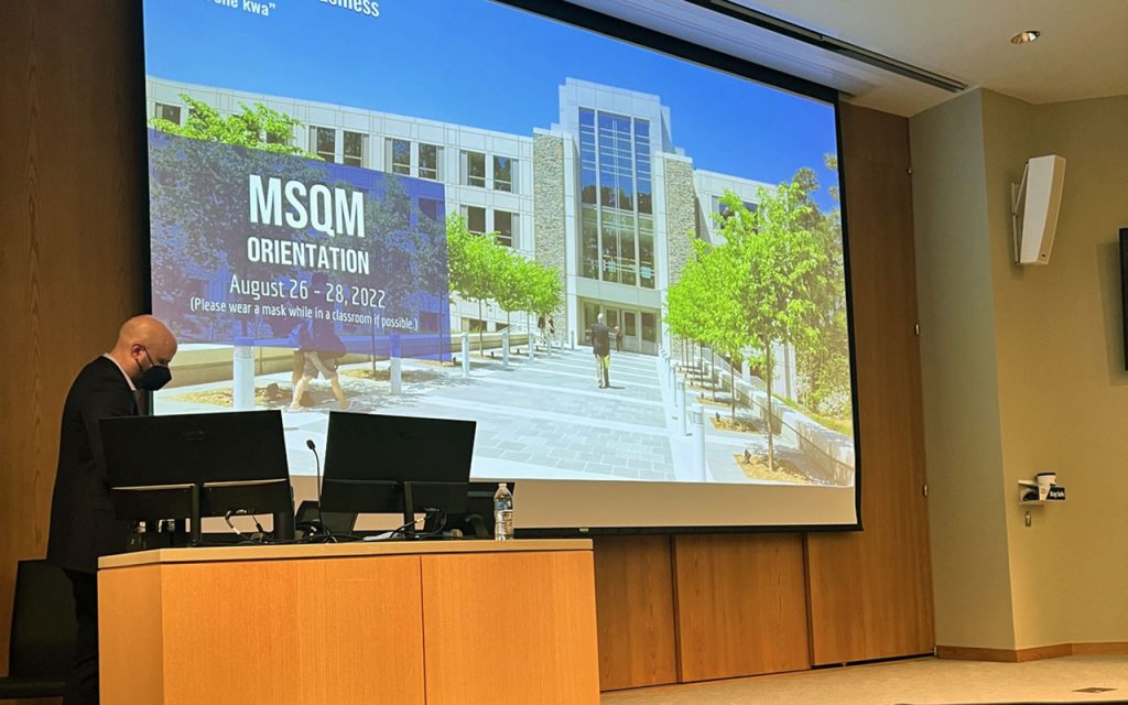 Senior Associate Dean Jeremy Petranka stands at a podium at the front of Geneen Auditorium at Duke's Fuqua School of Business. He is wearing a mask. a screen shows the a slide reading "MSQM orientation"