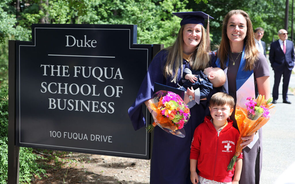 Jessica Meidinger, a graduate of the Weekend Executive MBA Class of 2025 at Duke University’s Fuqua School of Business, holds her young baby and a bouquet of flowers, standing with her wife and son next to a sign reading "Duke - The Fuqua School of Business - 100 Fuqua Drive"