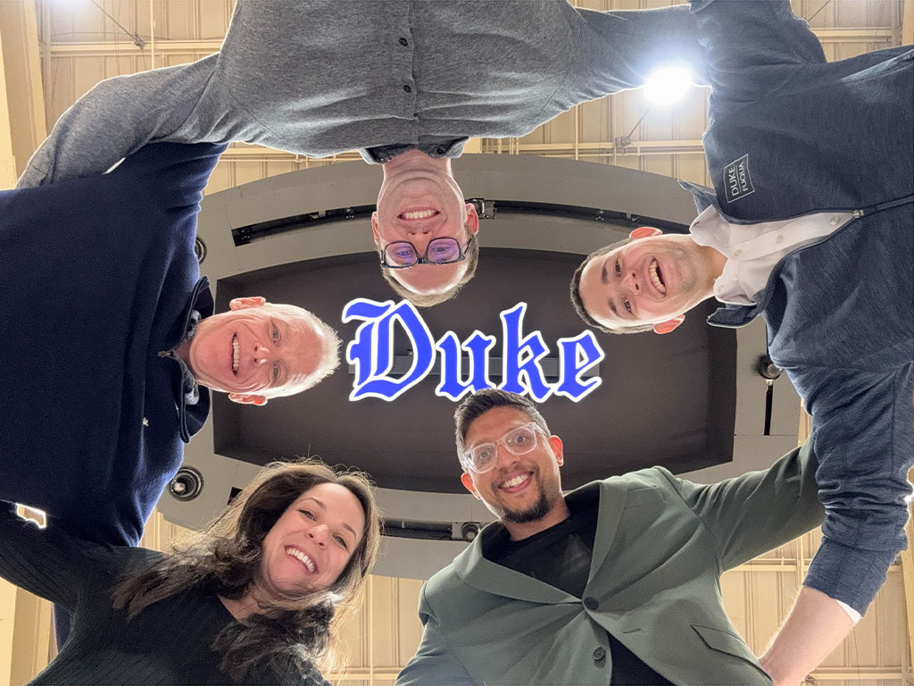 Ayan Bhandari with his classmates standing with their heads situated around a blue Duke logo on the jumbotron at Cameron Indoor Stadium high above them